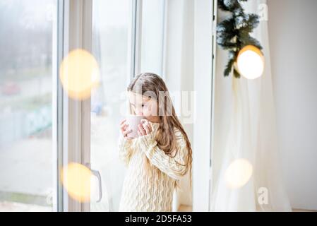 Ein kleines Mädchen in einem weißen Strickpullover trinkt heiße Schokolade, Kakao. Das Baby schaut aus dem Fenster, wartet auf ein Neujahrswunder Weihnachtsmann, isst Stockfoto