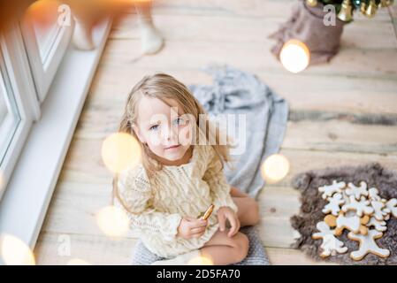 Ein kleines Mädchen in einem weißen Strickpullover trinkt heiße Schokolade, Kakao. Das Baby schaut aus dem Fenster, wartet auf ein Neujahrswunder Weihnachtsmann, isst Stockfoto