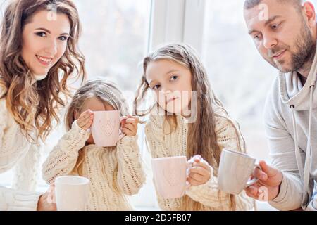 Glückliche Familie in weißen Pullovern trinkt Kakao am Panoramafenster. Eltern und Töchter halten Becher in den Händen, auf einem Teller sind Lebkuchen Stockfoto