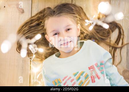 Glückliches Mädchen liegt auf dem Holzboden in ihren Haaren mit einer hellen Girlande von Weihnachtslichtern. Lächelnd, Blick auf die Kamera. Aufnahmen von oben, Stockfoto