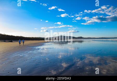 Zwei Personen spazieren entlang des tropischen Sandstrandes mit blauem Himmel und weißen Wolken, die sich im Wasser spiegeln, mit Bergen im Hintergrund Stockfoto