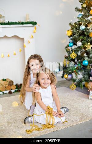 Zwei kleine blonde Mädchen in weißen Kleidern spielen am Kamin und am Weihnachtsbaum mit Kisten. Schwestern haben Spaß, umarmen, lachen, entfalten Geschenke Stockfoto