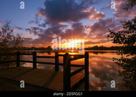 Erstaunlicher Sonnenaufgang in ländlicher Umgebung. Symmetrie des Himmels in einem See bei Sonnenuntergang. Wolken spiegeln sich auf dem Wasser. Holzponton im Vordergrund. Stockfoto