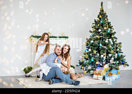 Eine glückliche Familie sitzt auf dem Teppich am weißen Kamin mit einem eleganten Weihnachtsbaum in Lichterketten. Junger Vater und schöne Mutter Stockfoto