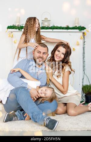 Eine glückliche Familie sitzt auf dem Teppich am weißen Kamin mit einem eleganten Weihnachtsbaum in Lichterketten. Junger Vater und schöne Mutter Stockfoto