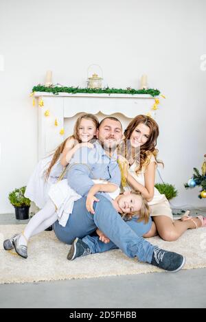 Eine glückliche Familie sitzt auf dem Teppich am weißen Kamin mit einem eleganten Weihnachtsbaum in Lichterketten. Junger Vater und schöne Mutter Stockfoto