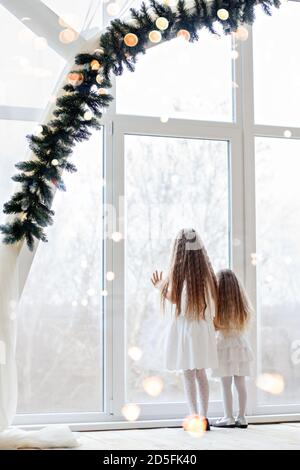 Zwei Freundinnen, Schwestern, blond in weißen Festkleider, blicken in das Panoramafenster mit Zweigen von Weihnachtsbäumen in Erwartung des Neuen Stockfoto