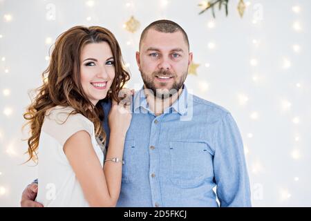 Ein liebevolles Paar hat Spaß auf dem Hintergrund einer weißen Wand mit Girlanden von hellen Lichtern, ein Zweig eines Weihnachtsbaums hängt von oben. Liebhaber glücklich Stockfoto