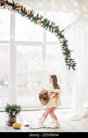 Zwei Freundinnen der blonden Schwester in gestrickten weißen Pullovern Und die Socken spielen am Panoramafenster mit Weihnachten Bäume in Erwartung Stockfoto