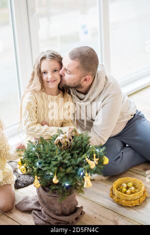 Liebevoller Vater küsst seine Tochter in der Nähe des Weihnachtsbaums. Ein junger Mann mit einem kleinen Mädchen schmückt einen Weihnachtsbaum in einem Topf mit goldenem Spielzeug Stockfoto