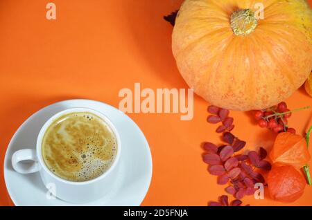 Weiße Tasse Kaffee mit Cappuccino-Creme auf einem orangen Hintergrund mit Herbstblättern, Äpfeln und roten Beeren Kürbis. Herbstsaison, Freizeit und Stockfoto