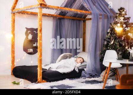 Ein kleiner Junge liegt bedeckt mit einer weißen Decke auf seinem Bett in Form eines Zelthauses, geschmückt mit Lichterketten. Frohe Kindheit. Kinder Stockfoto