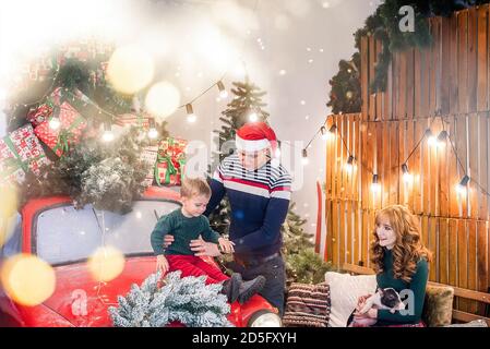 Eine glückliche Familie hat Spaß mit einem kleinen Sohn und einem hausgemachten Mini-Schwein in der Nähe eines roten Autos mit Geschenken, mit Weihnachtszweigen, Girlande, einem Kranz geschmückt Stockfoto