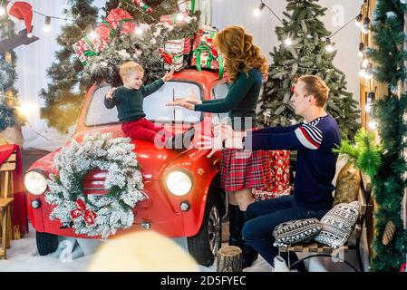 Eine glückliche Familie hat Spaß und spielt mit ihrem kleinen Sohn in der Nähe eines roten Autos mit Geschenken, geschmückt mit Weihnachtsbaum. Mutter und Vater umarmen den Jungen Stockfoto