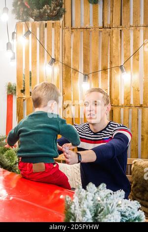 Ein junger Vater spielt mit seinem kleinen Sohn und einem Mini-Schwein vor einem roten Festwagen, geschmückt mit Lichterketten, Weihnachtsbäumen mit Bokeh. Stockfoto