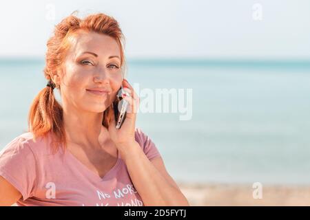 Nahaufnahme Porträt einer erwachsenen schönen Frau am Telefon anrufen. Im Hintergrund das Meer und ein Sandstrand. Speicherplatz kopieren. Konzept der Kommunikation Stockfoto
