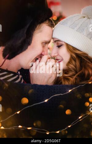 Ein Nahaufnahme-Porträt eines verliebten jungen Paares vor dem Hintergrund von Weihnachtsbäumen mit Girlanden. Ein junger Mann in einem Hut mit Ohrenklappen wärmt Stockfoto