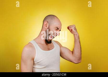 Menschen und Emotionen. Ein kahler, bärtiger Mann in einem weißen T-Shirt klatscht seine Faust vor Freude und schreit vor Freude. Gelber Hintergrund. Speicherplatz kopieren. Stockfoto