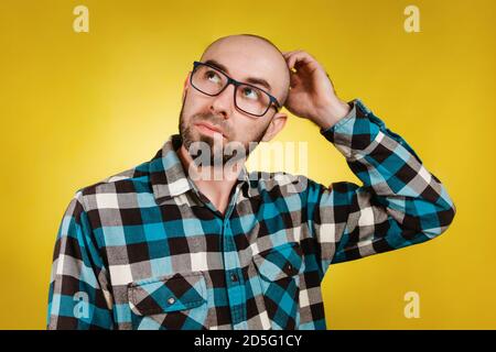 Menschen und Emotionen. Ein kahler, bärtiger Mann in einer Brille und einem blauen karierten Hemd, der seinen Kopf für eine Lösung oder eine Idee kratzt. Gelber Hintergrund. SPA kopieren Stockfoto
