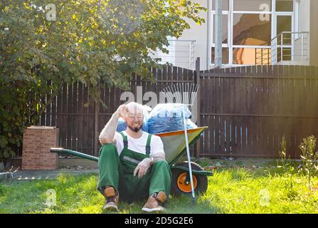 Der Gärtner in der grünen Uniform ruht auf dem Gras. Lächeln und gute Laune. Die Sonne scheint auf der rechten Seite. Stockfoto
