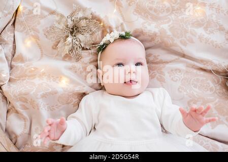 Nahaufnahme Porträt eines kleinen Babys in einem weißen Kleid mit einem Ornament auf dem Kopf. Das Mädchen liegt auf einem hellen Sofa in Girlanden von Lichtern, Weihnachtsbaum Stockfoto
