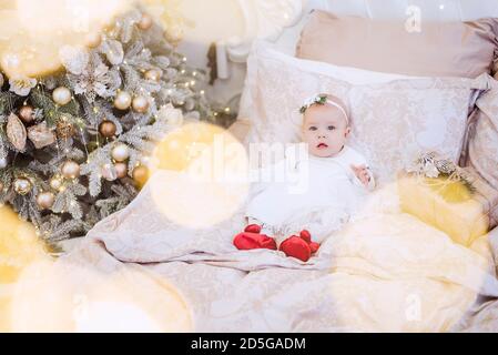 Nahaufnahme Porträt eines kleinen Babys in einem weißen Kleid mit einem Ornament auf dem Kopf. Das Mädchen liegt auf einem hellen Sofa in Girlanden von Lichtern, Weihnachtsbaum Stockfoto