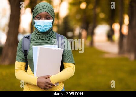 Schwarze muslimische Studentin trägt medizinische Maske im Freien, hält Rucksack und Bücher Stockfoto