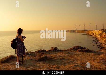 Eine schwangere Fotografin im floralen Sommerkleid und Rucksack macht ein Landschaftsfoto vom Windpark in Bozcaada, Türkei. Sie ist es Stockfoto