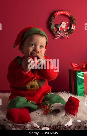 Süßer einjähriger Junge, gekleidet als Elfe, sitzend auf weißem Teppich und in Weihnachtsdekoration, mit Geschenken Stockfoto