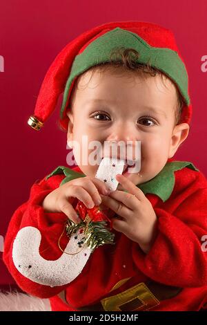 Süßer einjähriger Junge, der als Elfe sitzend gekleidet ist und einen typischen Weihnachtsstab im Mund hält Stockfoto