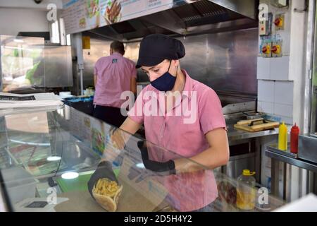 Junge Frau, die Essen (Gyros, Kebab) für einen Kunden vorbereitet, während sie eine Gesichtsmaske und Handschuhe trägt, aufgrund von Covid-19 Vorsichtsmaßnahmen Stockfoto