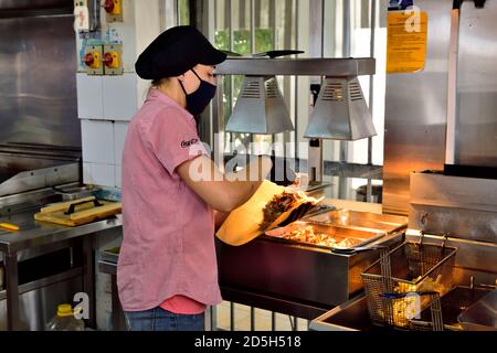 Junge Frau, die Essen (Gyros, Kebab) für einen Kunden vorbereitet, während sie eine Gesichtsmaske und Handschuhe trägt, aufgrund von Covid-19 Vorsichtsmaßnahmen Stockfoto