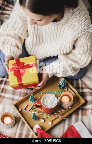 Junge Frau sitzt auf dem Bett in kuschelig gestrickten Wollpullover und hält Geschenkbox mit Rotgold. Hygge, Neujahr, Weihnachten. Holztablett mit einem Becher Stockfoto