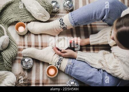 Junge Frau sitzt auf karierten in kuschelig gestrickten weißen Wollpullover und Socken hält eine Tasse Kakao in den Händen. Hygge Neujahr, gemütliche Weihnachten, Vorbereitung Stockfoto