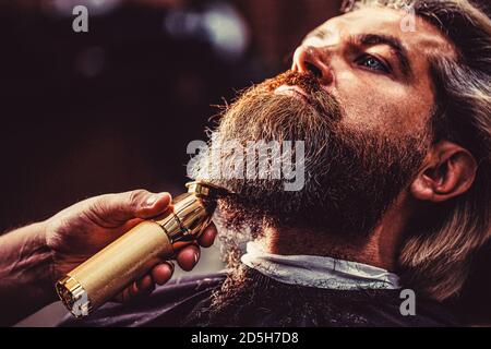 Bärtiger Mann im Friseurladen. Mann, der Friseur im Friseursalon besucht. Barber arbeitet mit einem Bartschneider. Hipster-Client immer Frisur Stockfoto