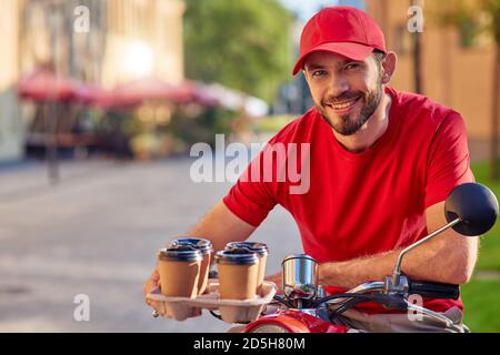 Junger kaukasischer männlicher Kurier lächelt an der Kamera, während er vier Kaffeetassen auf dem Roller entlang der sonnigen Straße liefert. Konzept für die Lebensmittelzustellung Stockfoto