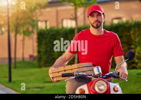 Junge gut aussehende kaukasischen Delivery Mann in Uniform auf Roller halten Pizzakisten. Konzept für die Lebensmittelzustellung Stockfoto