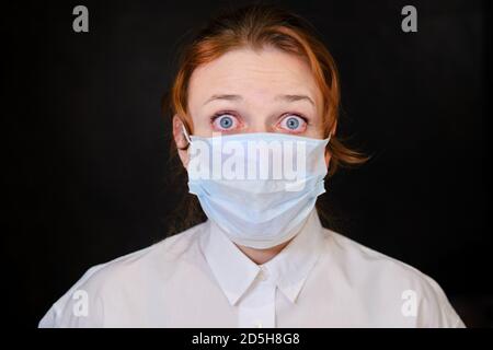 Eine junge Frau mit verängstigten Augen in einer medizinischen Maske auf schwarzem Hintergrund, Konzept. Rothaarige Mädchen während der Coronavirus Quarantäne Nahaufnahme Stockfoto