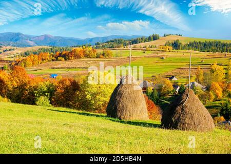 Ländliche Landschaft in den Bergen. Heuhaufen auf dem Hügel. Dorf im Tal. Landschaft in Herbstfarben. Schönes sonniges Wetter mit flauschigen Wolken auf den s Stockfoto