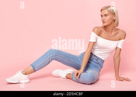 Freistil. Junge blonde Frau in Jeans und oben sitzend isoliert auf rosa Blick Seite neugierig Stockfoto
