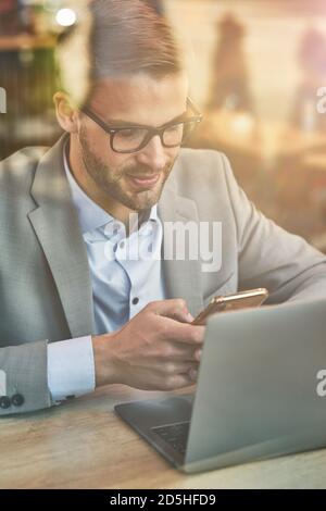 Chatten mit Freunden. Vertikale Aufnahme eines jungen gutaussehenden Geschäftsmannes oder männlichen Freiberuflers mit seinem Smartphone während der Online-Arbeit im Café, Laptop auf Stockfoto