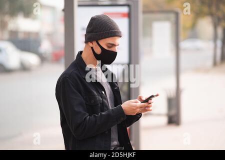 Mann in Maske mit Handy an einer Bushaltestelle. Millennial mit Smartphone am öffentlichen Ort, covid Zeit Stockfoto