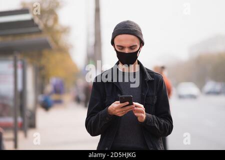 Mann in Maske mit Handy an einer Bushaltestelle. Millennial mit Smartphone am öffentlichen Ort, covid Zeit Stockfoto