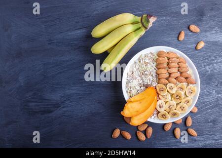 Ein Teller mit Müsli, Mandeln, Banane, Persimmon auf schwarzem Holzgrund. Draufsicht. Kopierraum Stockfoto