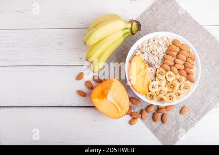 Ein Teller mit Müsli, Mandeln, Banane, Persimmon auf weißem Holzgrund. Draufsicht. Kopierraum Stockfoto