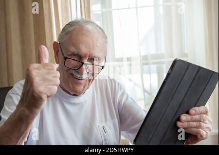 Glücklicher älterer Mann zu Hause mit Laptop. Ein grauhaariger alter Mann mit Brille ist glücklich und streicht, dass alles in Ordnung ist. Das Konzept der Kommunikation Stockfoto