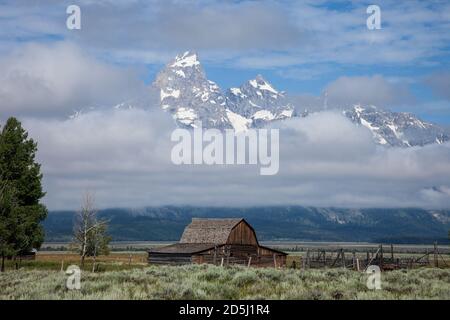 Die alte Holzscheune auf dem John Moulton Gehöft in der Mormon Row im Grand Teton National Park mit dem Grand Teton, der aus den Wolken dahinter guckt. Wyom Stockfoto