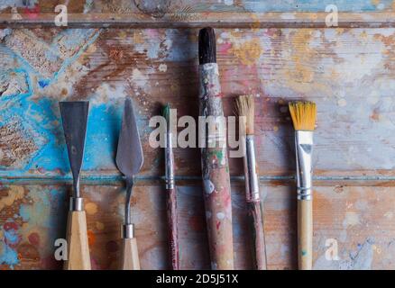 Künstlerschreibtisch und Materialien.verschiedene Pinsel & Pallete Messer.der Holzschreibtisch ist mit schönen verschiedenen Farben bedeckt. Stockfoto