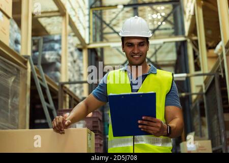 Junge indische Industriefabrik Lagerarbeiter arbeiten in der Logistikindustrie indoor. Lächelnder glücklicher Mann, der eine Zwischenablage hält und Artikel im Lager überprüft Stockfoto