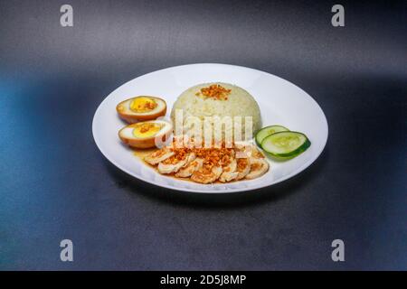 Hainanese Hühnerreis oder gedämpfter Hühnerreis mit süßem Sojasauce-Ei und Gurkenscheiben. Serviert auf einem weißen Teller. Stockfoto